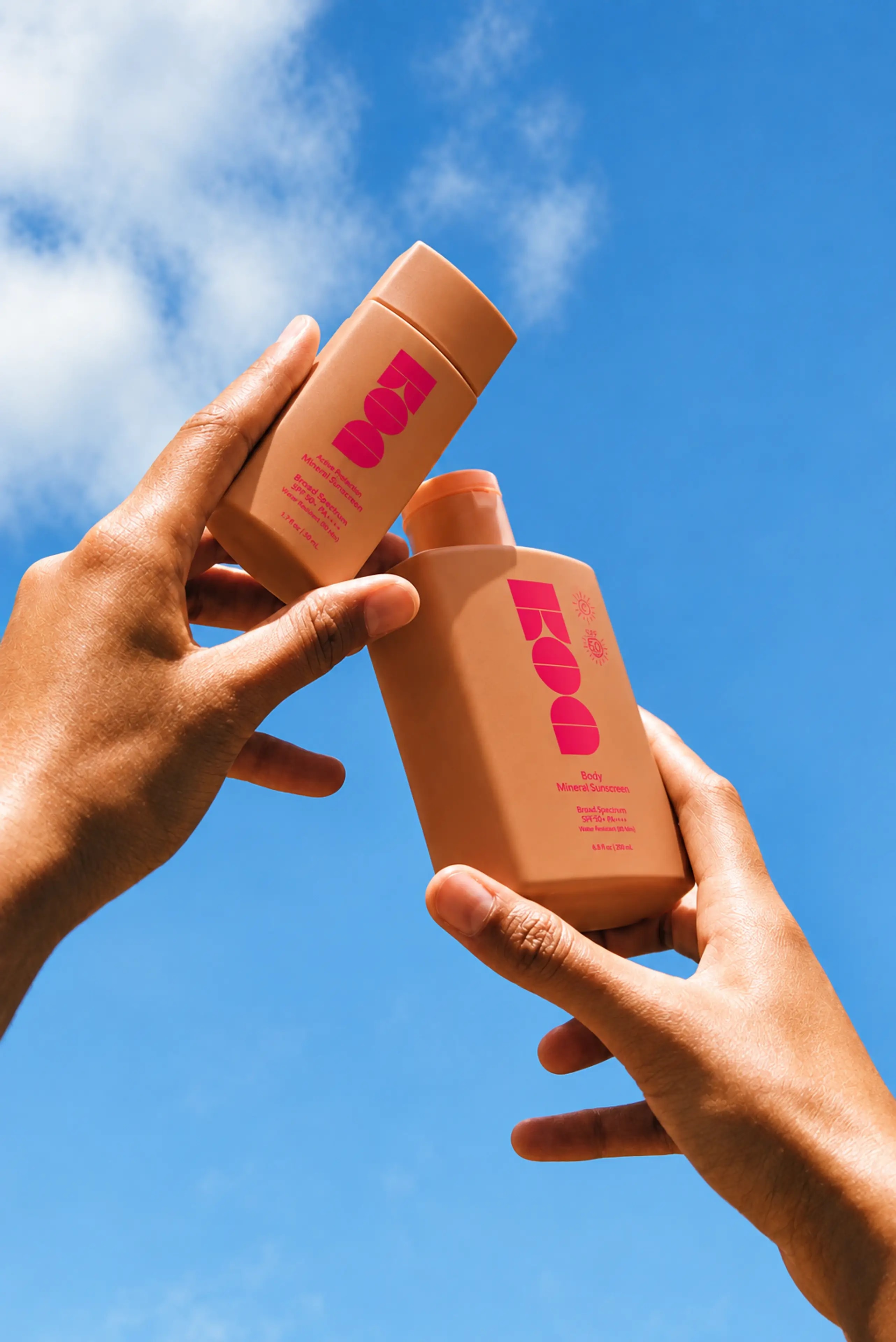 Two hands raising peach-colored bottles against a blue summer sky
